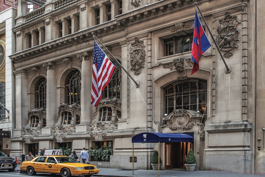 A yellow taxi sits parked outside an old-fashioned light brown building with two flags hanging outside.