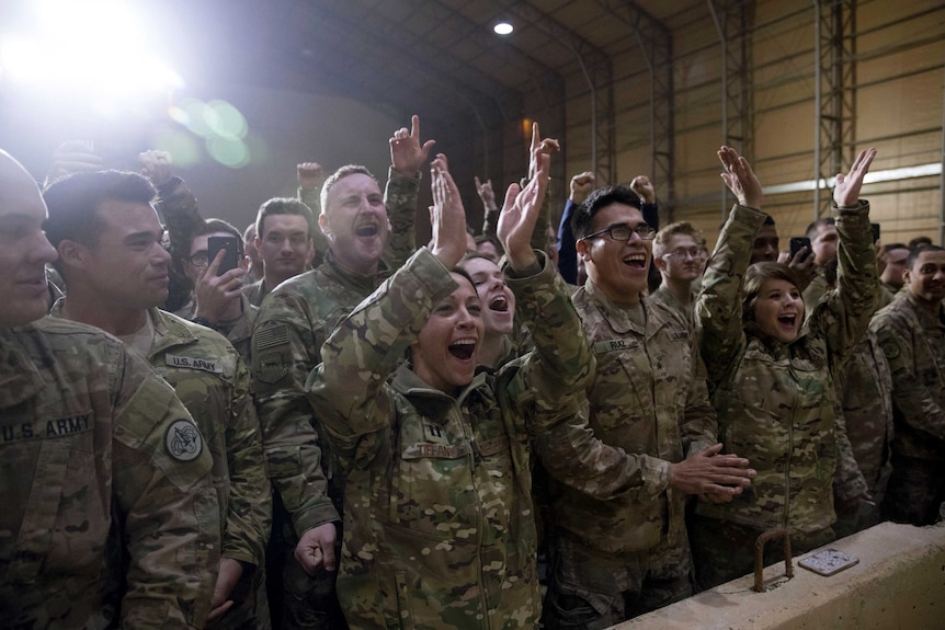 Rows of US military officers cheer behind a concrete barricade in an air hangar as a camera flash fires behind them.