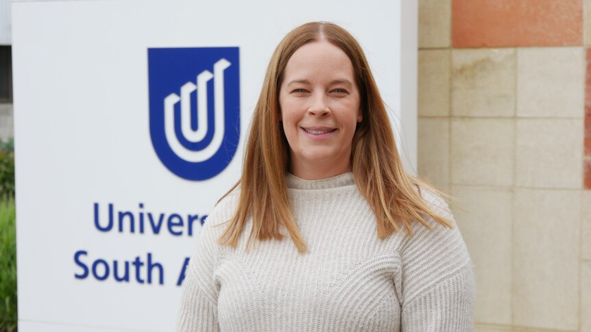 A smiling woman with long ginger hair standing in front of a sign that reads "University of South Australia".