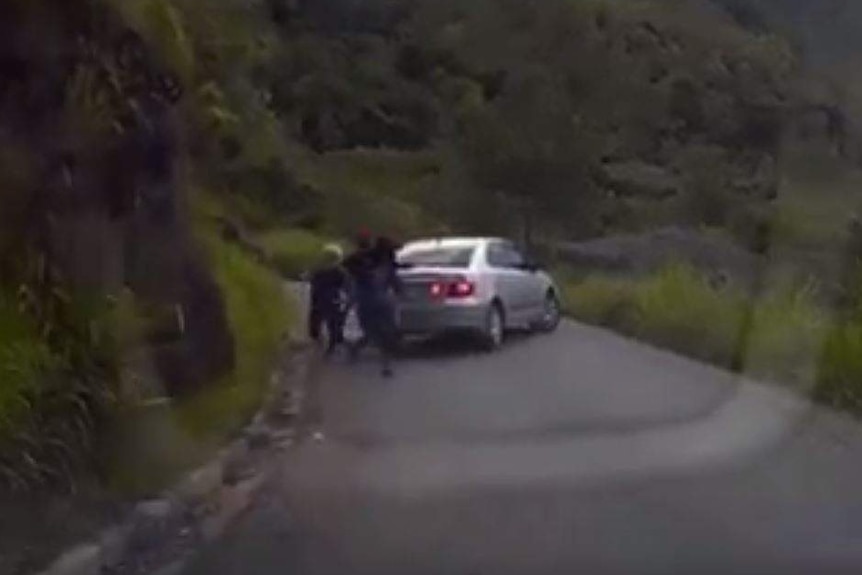 A silver car blocks a narrow road in Papua New Guinea and three dark figures leave the car to chase an Australian family.