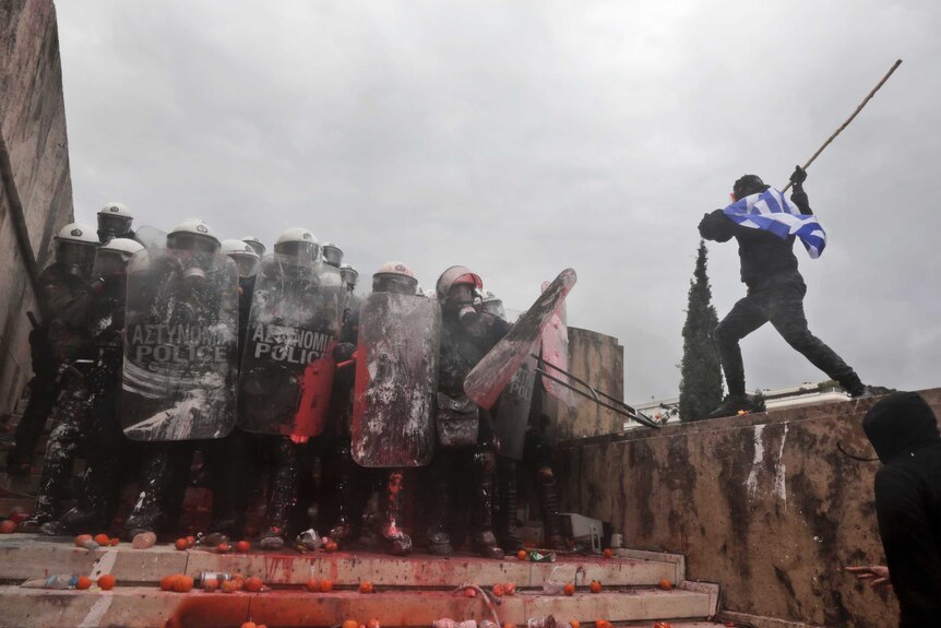 Look up a stone steps, a protester draped in the Greek flag attacks riot police from an adjacent ledge with a stick.