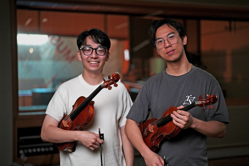 Brett Yang and Eddy Chen standing together holding a violin each, studio behind.