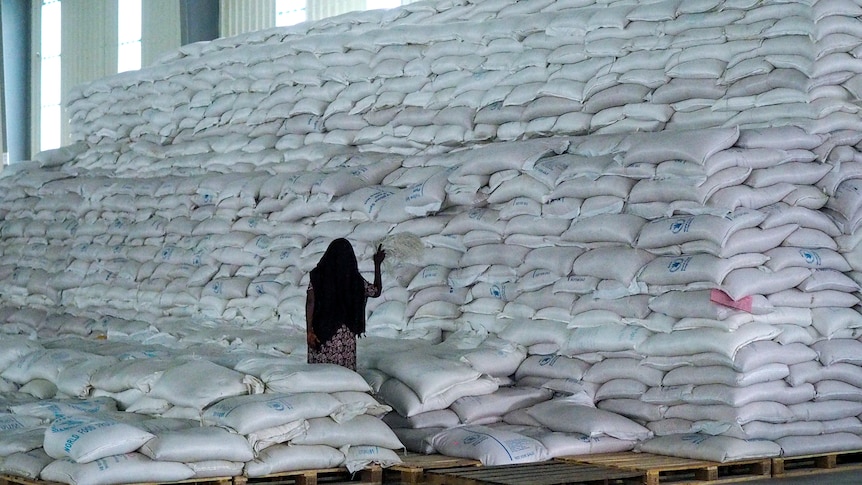 A woman in a black shawl tends to a massive pile of white sacks in a well-lit warehouse.