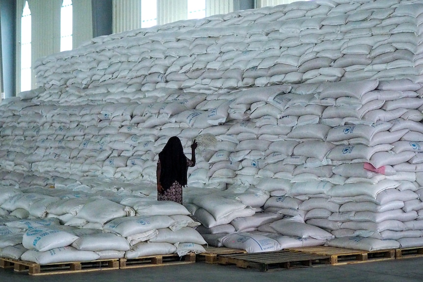 A woman in a black shawl tends to a massive pile of white sacks in a well-lit warehouse.