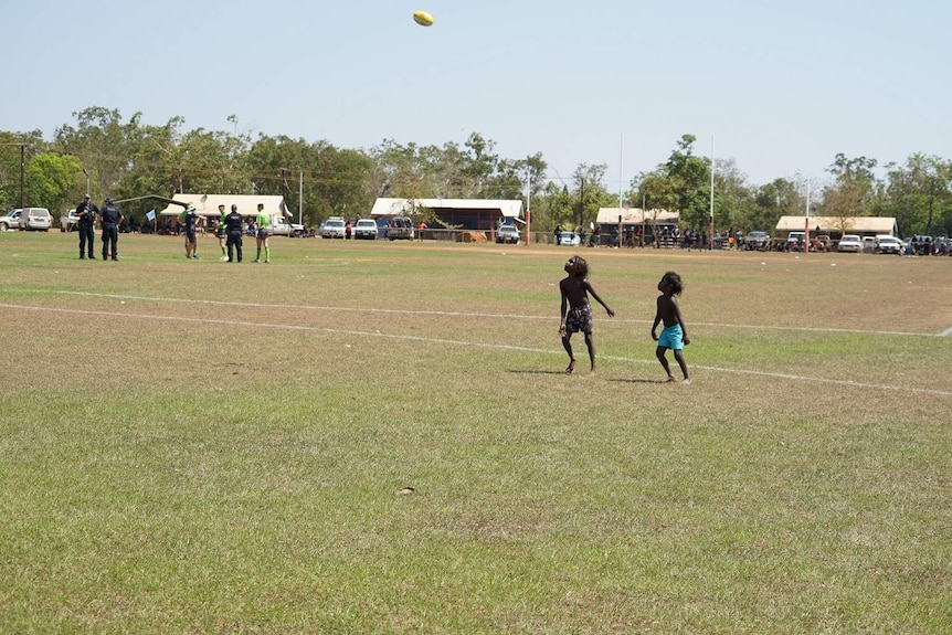 Two kids kicks the footy on the Wadeye oval as umpires and police stand in the centre of the ground.