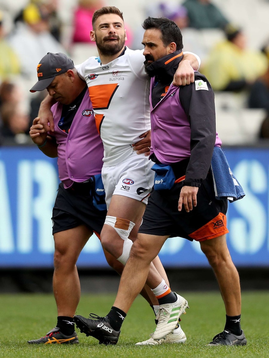A male AFL player looks to his left as he is helped from the field injured with his arms around two medical staff members.