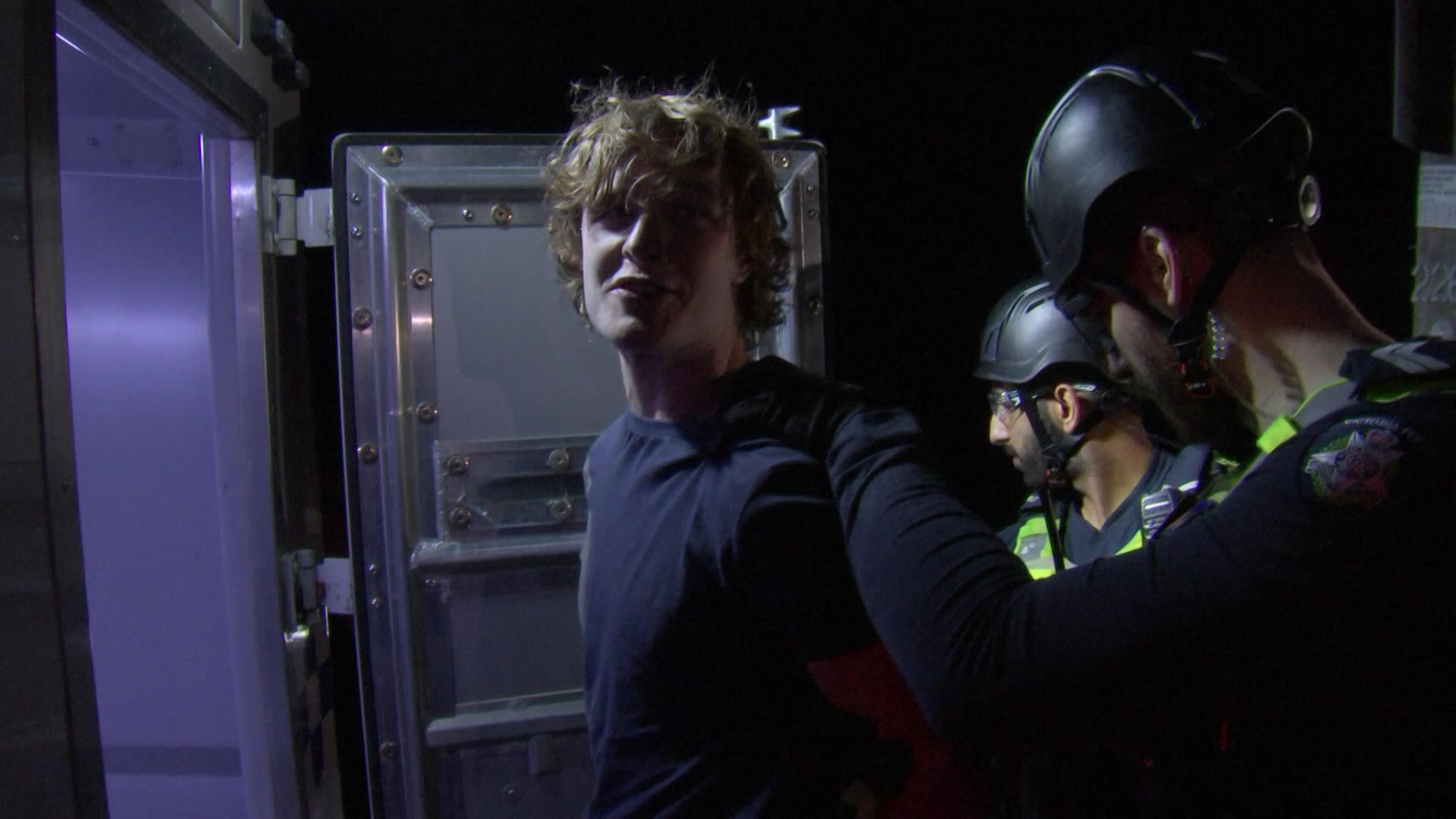 A man being loaded into a police van by two officers wearing helmets.