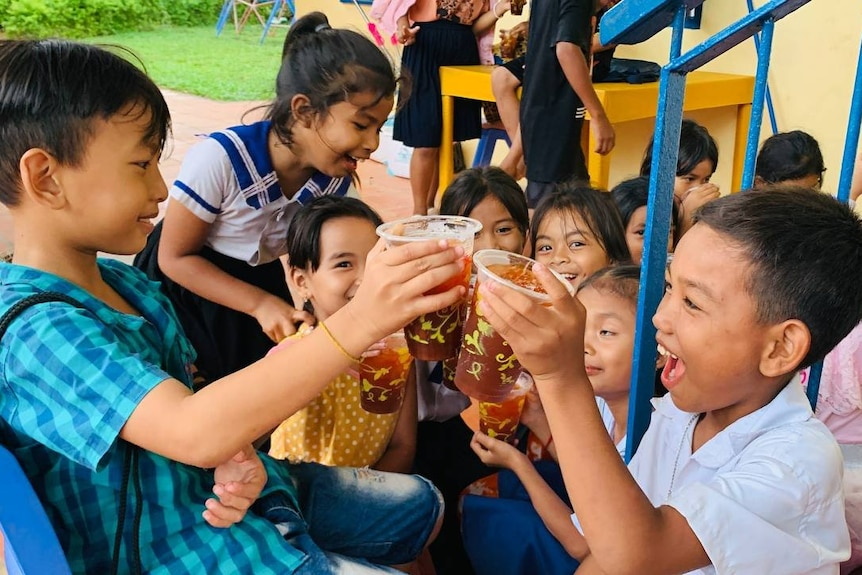 two children toasting drinks in plastic cups with other children smiling and playing in background