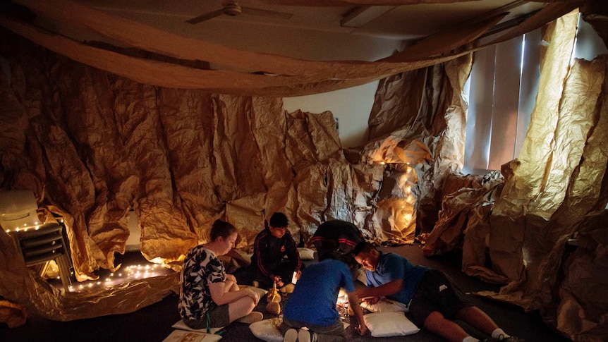 A group of kids and a teacher sit in a lit-up papered room