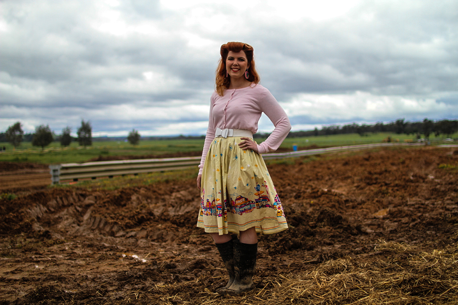 A woman in gumboots, flared skirt and pink cardigan stands in the middle of a muddy field.