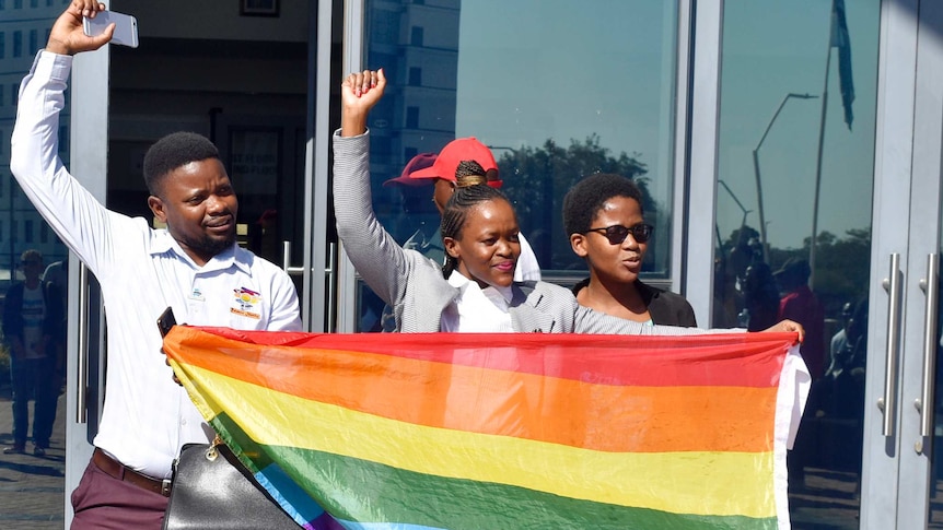 Three LBGTI activists hold the rainbow flag and raise their arms in celebration outside court