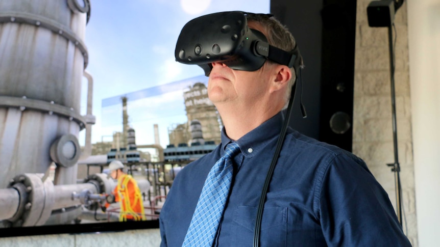 Doug Bester wears a 3D iPhone virtual reality mask. A worker in high viz works in the background.