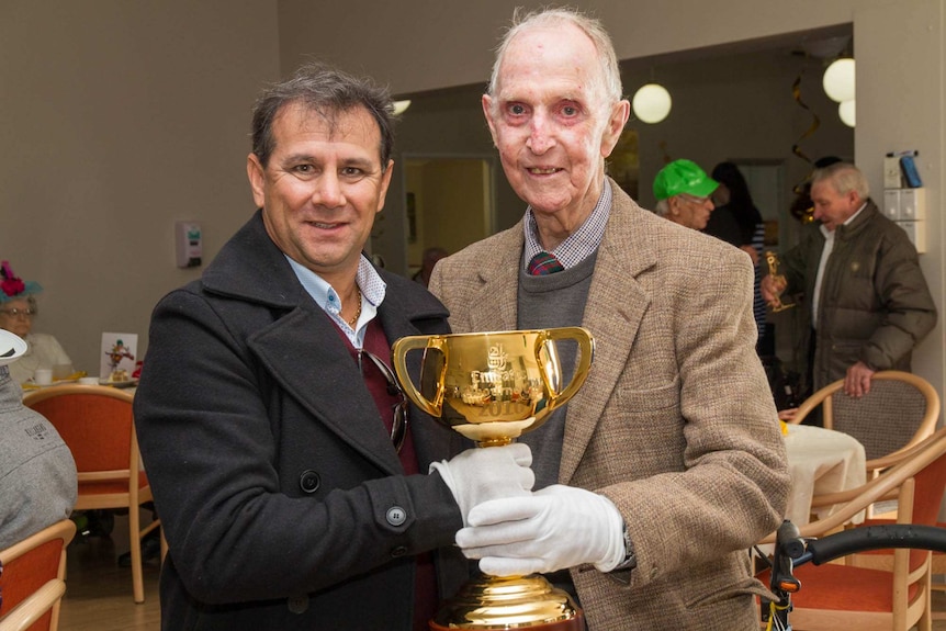 Two men wearing white gloves holding a gold cup labelled Emirites Melbourne Cup 2016