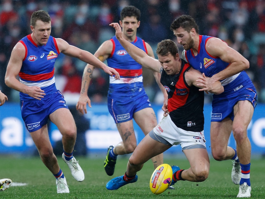 An Essendon AFL player puts his arms out as he falls to ground near the ball as he is surrounded by Western Bulldogs players.