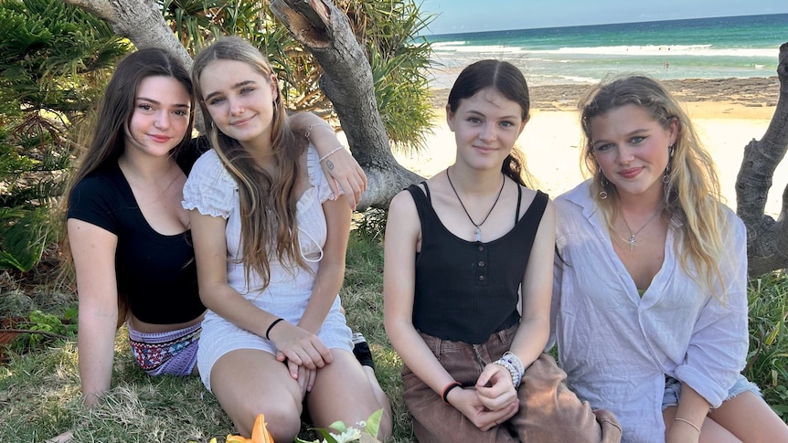 Four young women and girls sit together under a tree with a beach in the background.