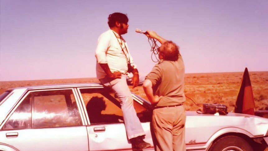 An Aboriginal man sits on top of a car being interviewed by a non Indigenous man with a microphone.