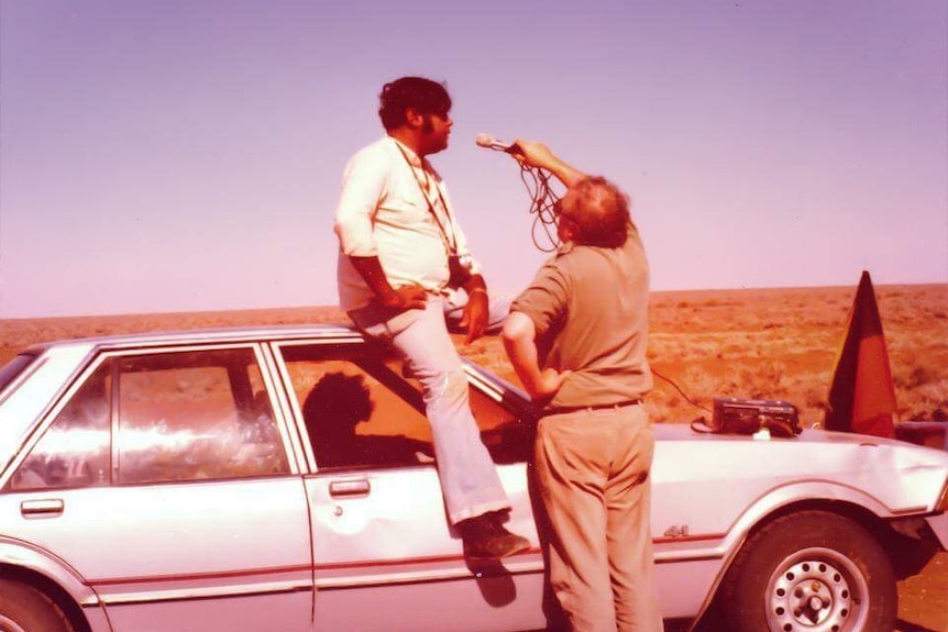 An Aboriginal man sits on top of a car being interviewed by a non Indigenous man with a microphone.