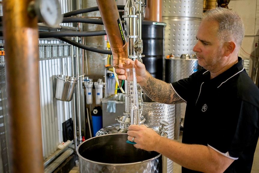 Man with greying hair pours alcohol from a large metal gin still.