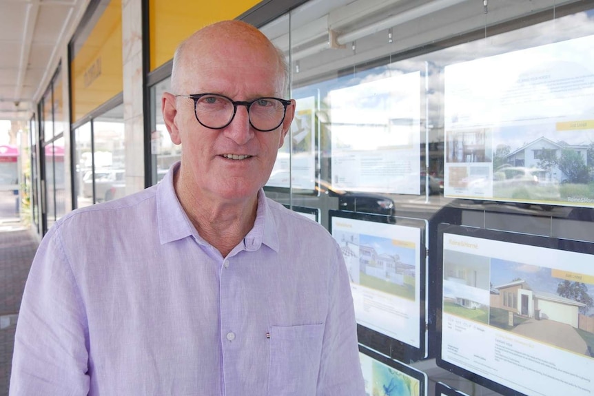 A balding, older caucasian man stands on a footpath outside a real estate agency.