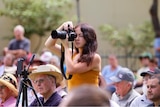 A woman stands above a seated crowd, holding an SLR camera to take a photo.