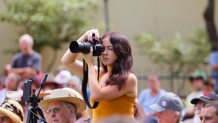 A woman stands above a seated crowd, holding an SLR camera to take a photo.