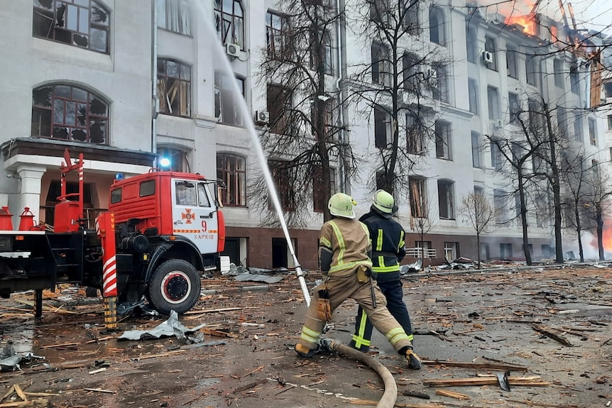 Firefighters spray water from a hose into the air as a building burns and rubble covers the ground