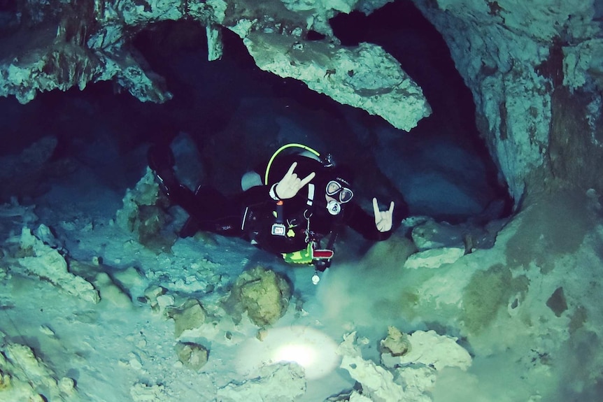 Man in wetsuit and scuba mask gestures hang loose with his hands, and is framed inside a cave