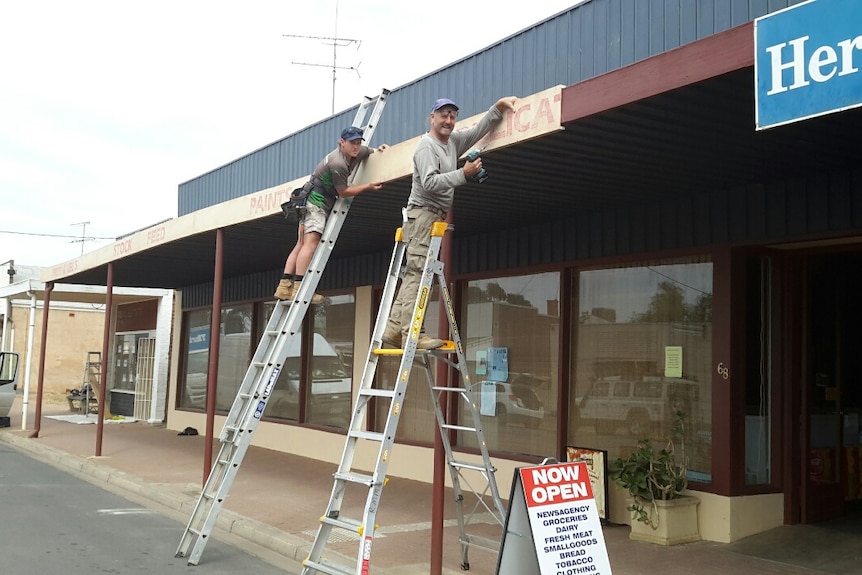 Two men stand on ladders. One his holding a drill and the other is helping hold the sign that's on the building's roof
