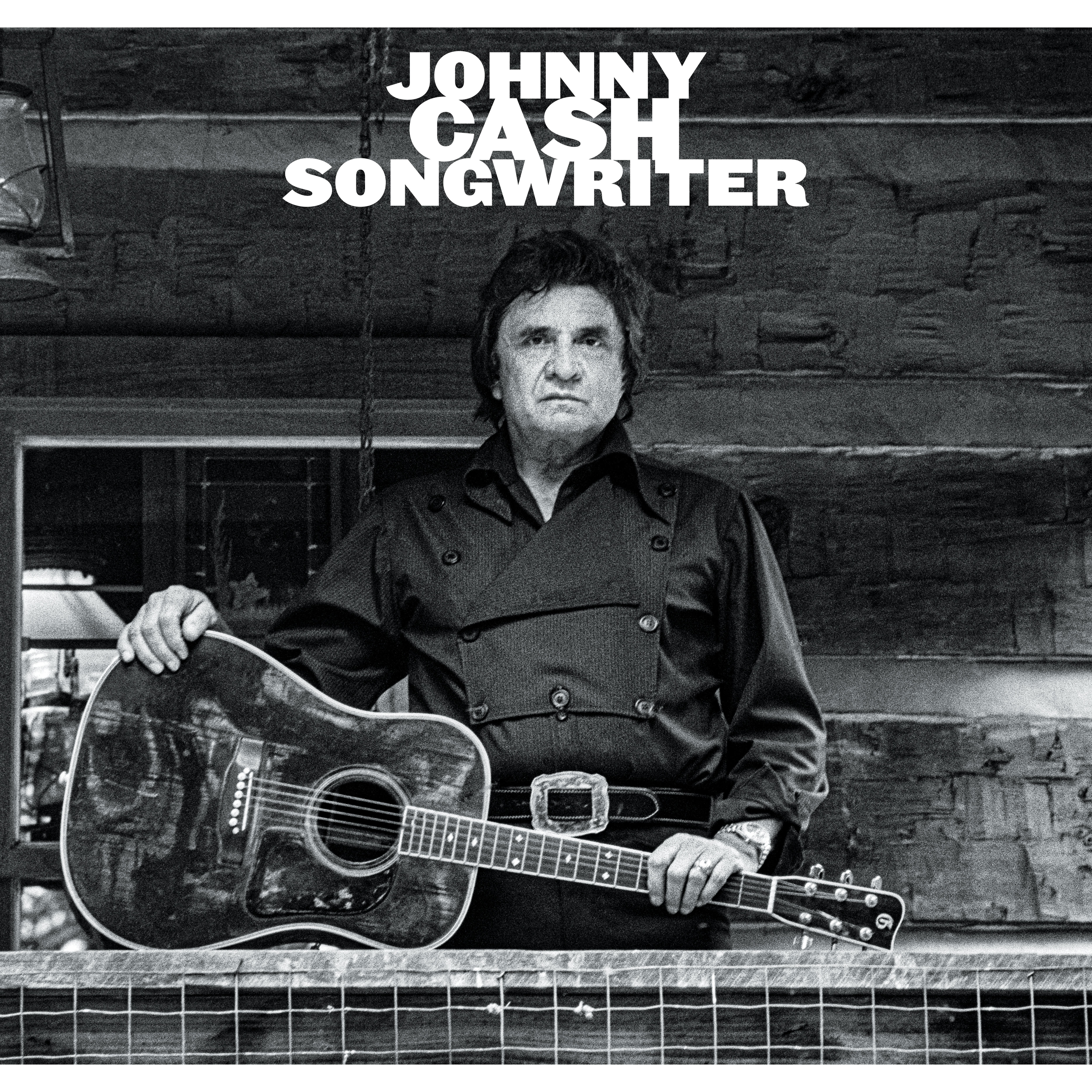 a stern faced johnny cash stands on the porch of a wooden building with an acoustic guitar