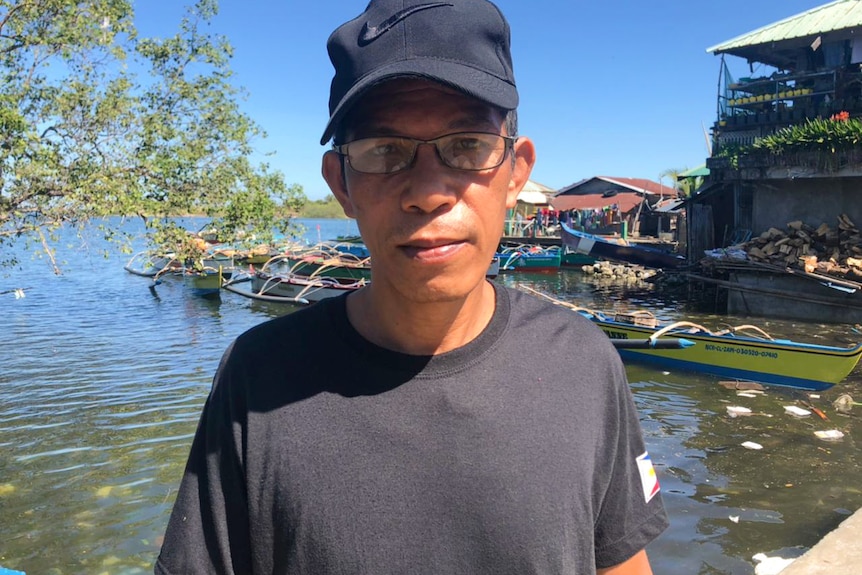 A man wearing glasses and a black cap and a black shirt stands in front of water.