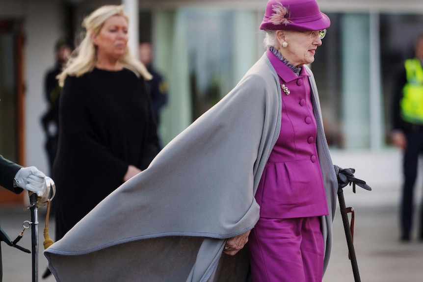 Queen Margrethe in a purple pant suit, matching hat and grey cape that is billowing in the wind