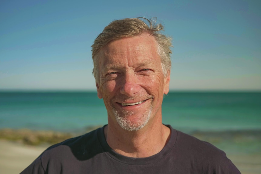 A close-up of a man with a light white goatee, squinting in the sun and wearing a black t-shirt with the ocean in the back
