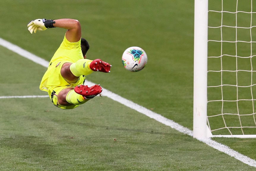 An A-League goalkeeper flings himself through the air to his right to stop a shot.