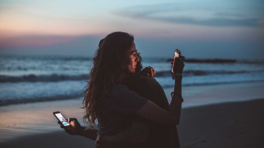 A man and woman hug each other while looking at their phones