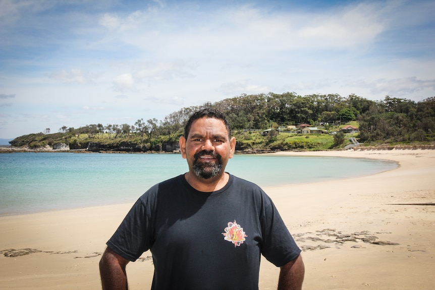 A bearded Indigenous man in a dark shirt stands on a beach with his hands on his hips.