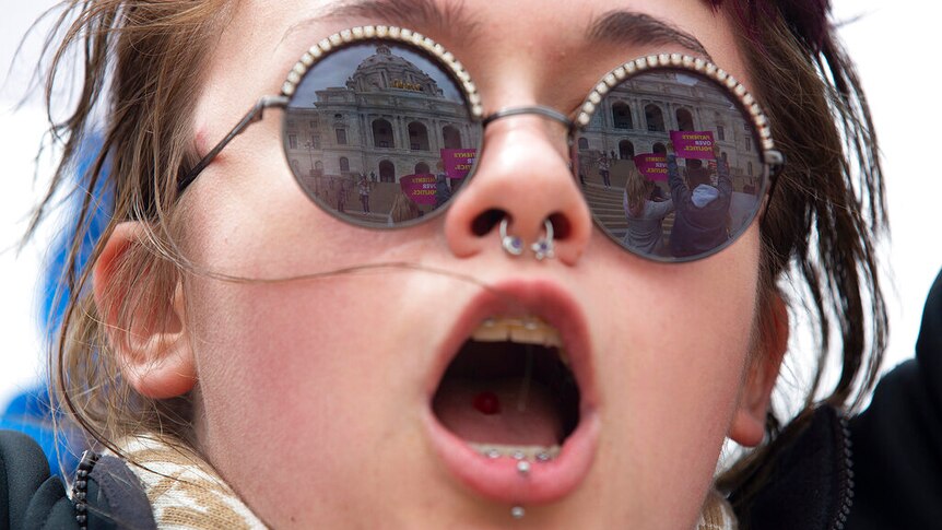 A young woman opens her mouth to shout during a protest, while other protesters and their banners are reflected in her glasses.