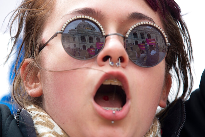 A young woman opens her mouth to shout during a protest, while other protesters and their banners are reflected in her glasses.