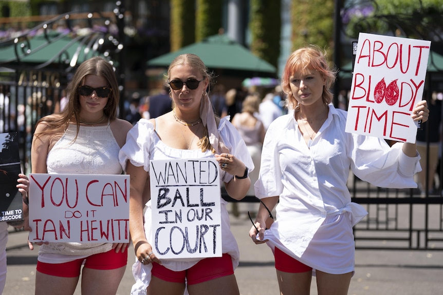 Three women in white shirts and red shorts hold signs. One reads: "About Bloody Time" with 'O's represented by blood droplets.