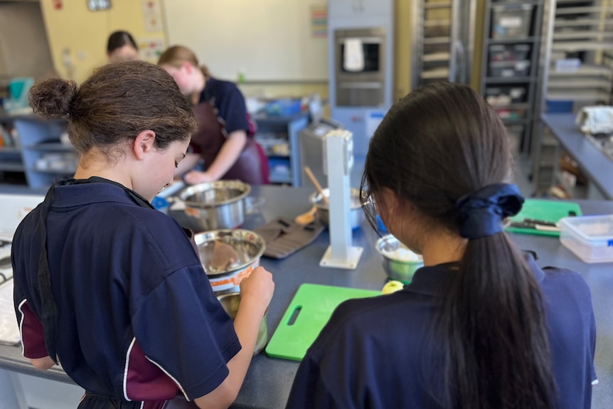 two year-7 female students in school uniform mixing ingredients in cooking class, with their backs facing the camera