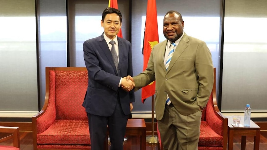 China's ambassador to PNG Xue Bing shakes hands with PNG Prime Minister James Marape in front of chairs and the PNG flag