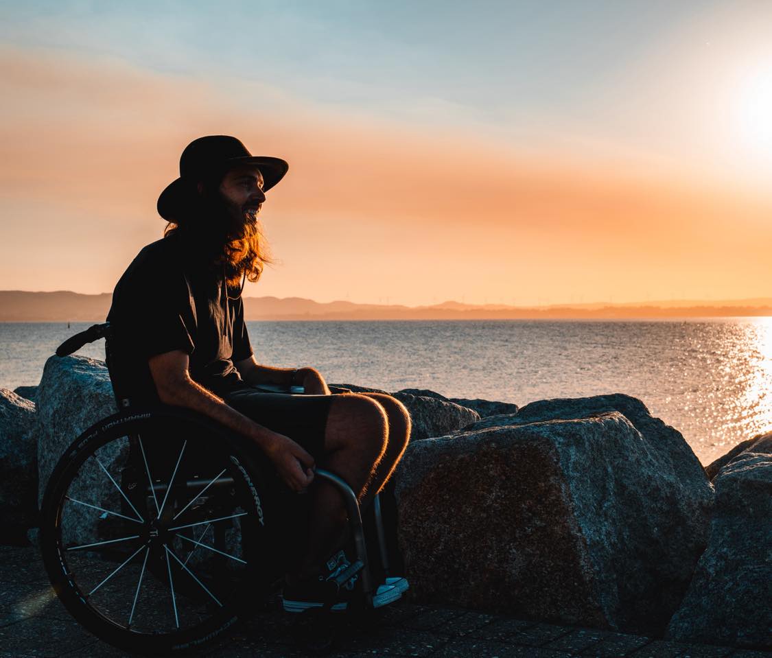 He sits in a wheelchair at a beach at sunset