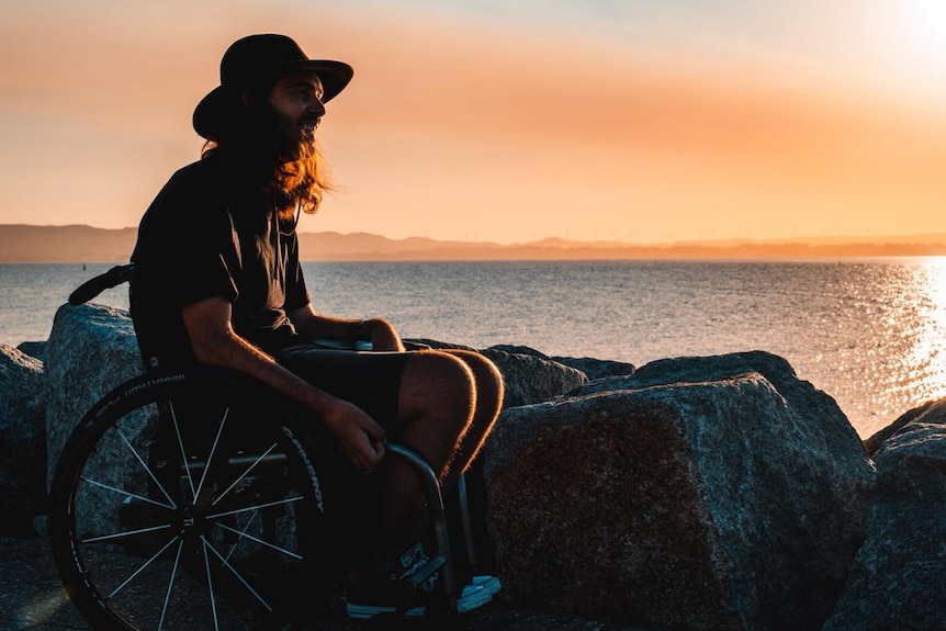 He sits in a wheelchair at a beach at sunset
