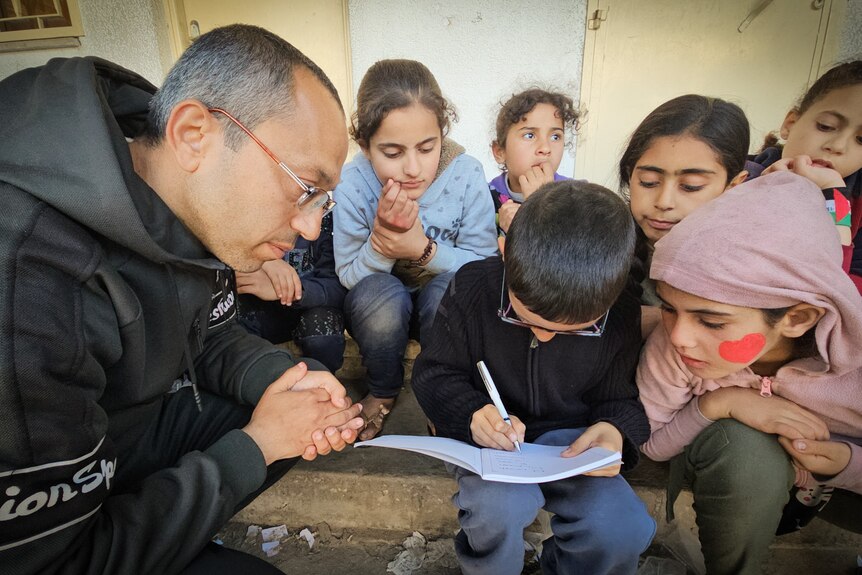 A man wearing a black hoodie looks down as a child writes on a piece of paper while other children watch.