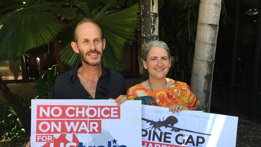 A man and woman standing under a tree holding Pine Gap protest signs