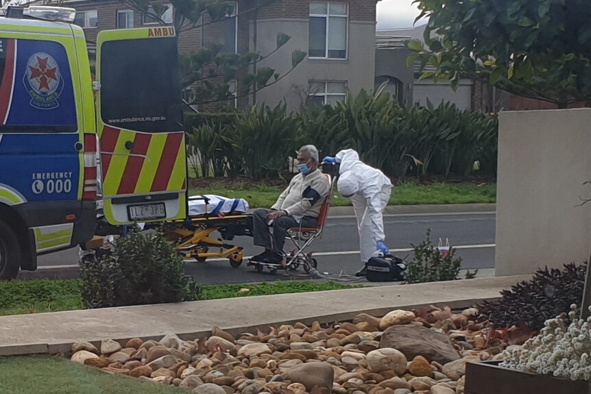 A man wearing a mask and sitting in a wheelchair is wheeled to the back of an ambulance by a paramedic in full PPE gear.