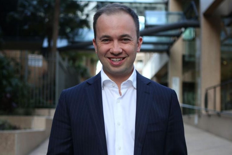 A man in a navy suit jacket and white-collared shirt smiles.