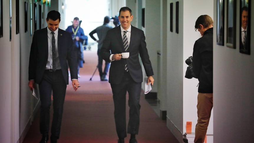 A man in a dark suit walks down a corridor holding a coffee cup with another man beside him.