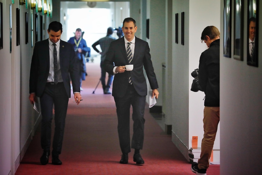 A man in a dark suit walks down a corridor holding a coffee cup with another man beside him.