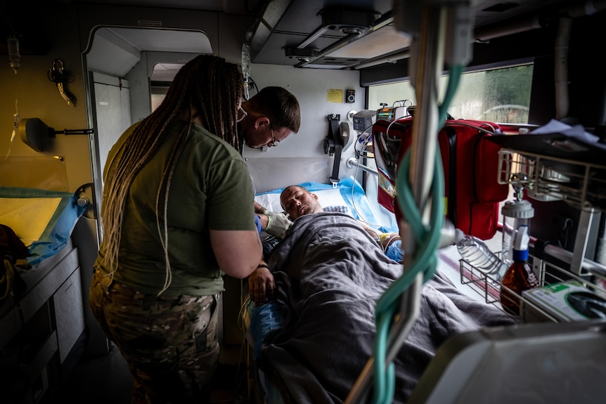 A man lays in a hospital bed inside a bus, while two medics stand nearby looking over him