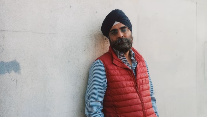 A South Asian man leans against a rough concrete wall, while wearing a navy turban, red puffer vest, and blue long-sleeve shirt.